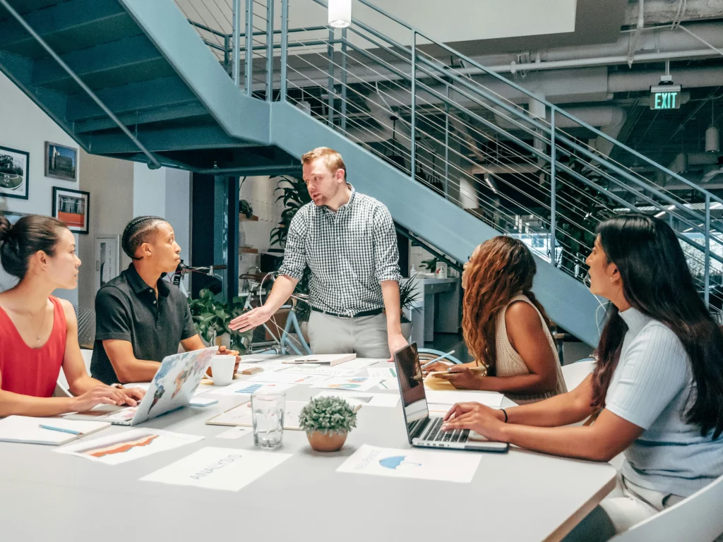 A group of people sat at a desk listening to their manager who is stood up at the head of the table