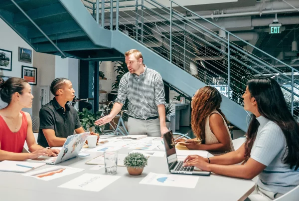 A group of people sat at a desk listening to their manager who is stood up at the head of the table