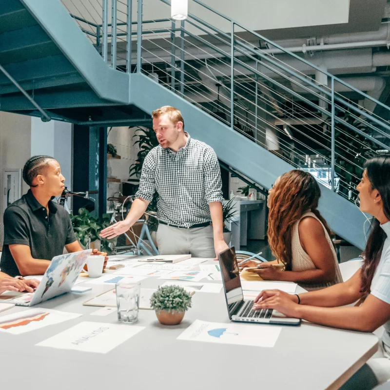 A group of people sat at a desk listening to their manager who is stood up at the head of the table