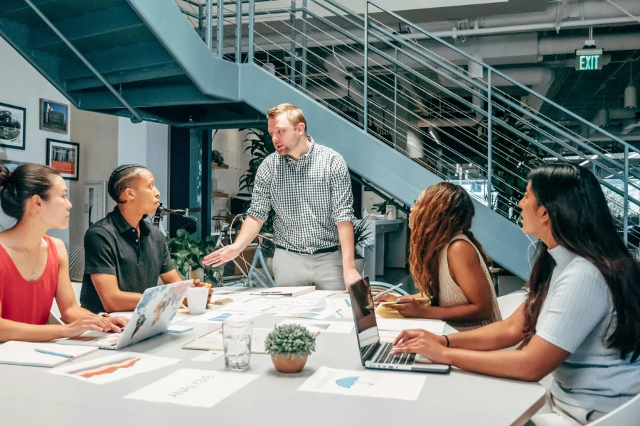 A group of people sat at a desk listening to their manager who is stood up at the head of the table